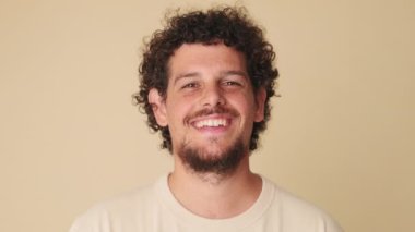 Yakından, genç adam kameraya bakıyor ve stüdyodaki bej arka planda izole bir şekilde gülüyor.