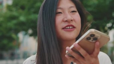 Kapalı, neşeli genç bir kadın şehir parkında bankta oturuyor ve hoparlörden konuşuyor.