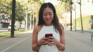 Gülümseyen genç bir kadın sokakta yürüyor, sosyal ağlarda cep telefonuyla iletişim kuruyor.