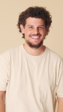 Mutlu genç adam kameraya bakarak gülüyor. Stüdyonun bej arka planında izole edilmiş.