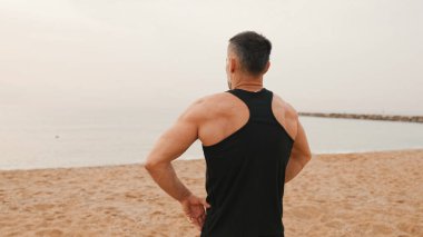 Olgun bir adam plajda dikilirken antrenmandan önce ısınıyor.