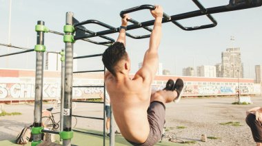 Guy, yatay bir çubuktaki sporcu bacaklarını kaldırır, yatay bir çubuğa asılır ve bir yaz günü açık hava spor sahasında karın kaslarını çalıştırır.