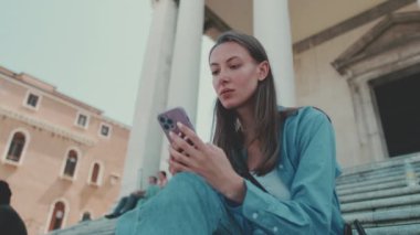 Uzun kahverengi saçlı, mavi gömlekli, Venedik 'te merdivenlerde otururken cep telefonu kullanan hoş bir kız.