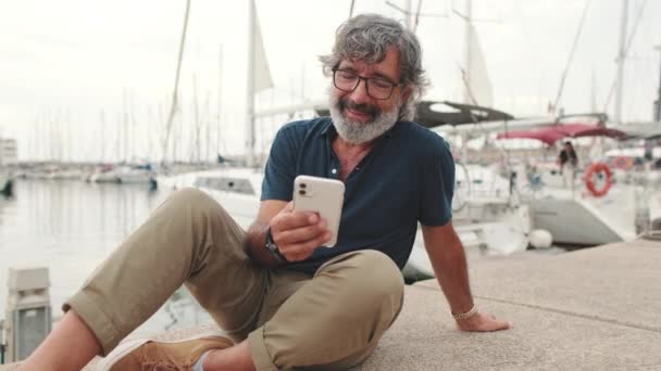 用电话微笑坐在港口的中年男人 — 图库视频影像
