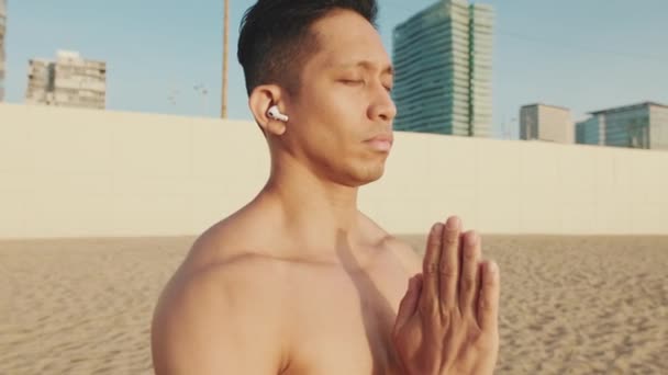 年轻的运动员戴着耳机站在海滩上做呼吸练习 — 图库视频影像