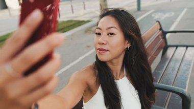 Beyaz bluzlu güzel esmer kız gülümsüyor ve kentin arka planında bankta otururken selfie çekiyor.