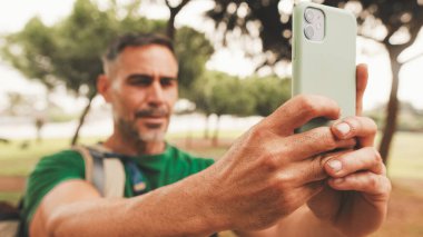 Omzunda sırt çantası olan orta yaşlı bir adam şehir parkında dikilirken cep telefonuyla fotoğraf çekiyor.