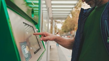 Günlük kıyafetler giyen olgun bir adam, toplu taşıma durağında dururken elektronik terminalden bilet alır.