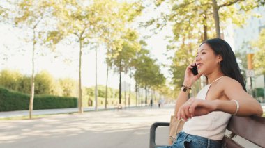 Beyaz bluz ve kot giyen tatlı esmer kız, kentin arka planında bankta oturuyor, akıllı telefondan konuşuyor.