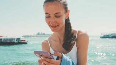 Uzun kahverengi saçlı, mavi gömlekli güzel bir kız, Venedik 'te deniz kenarında oturur ve arka planda cep telefonu, tekne ve sürat teknesi kullanır.