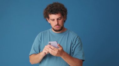 Elinde telefonla stüdyoda mavi arka planda baş parmak gösteren şaşırmış bir adam.