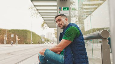 Sıradan kıyafetler giymiş orta yaşlı bir adam, şehirde toplu taşıma durağında tramvay beklerken cep telefonu kullanıyor.