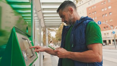 Günlük kıyafetler giyen olgun bir adam, toplu taşıma durağında dururken elektronik terminalden bilet alır.