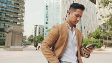 Bej ceketli genç işadamı elinde cep telefonuyla caddede yürüyor.