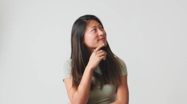 Gri elbiseli bir kız, bir konuda derin düşüncelere dalmış, gülümseyerek hayal kurmaya çalışıyor, beyaz arka planda izole edilmiş bir çeneye el desteği koyuyor, stüdyo çekimi.