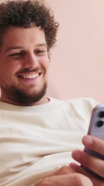 Oturma odasında kanepede oturan gülümseyen adam telefon ekranında kayıyor.