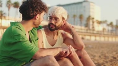 Homoseksüel çift kumsalda otururken kollarını kavuşturup sarılıyorlar.