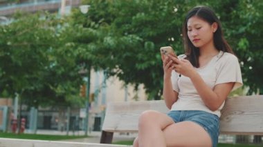 Şehir parkında bankta oturan genç bir kadın, cep telefonu kullanıyor.