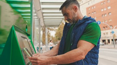 Günlük kıyafetler giyen olgun bir adam, toplu taşıma durağında dururken elektronik terminalden bilet alır.