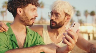 Yakın plan, sahilde otururken cep telefonu kullanan homoseksüel çift.
