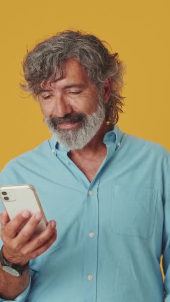 Ευτυχισμένος Ηλικιωμένος Γκριζομάλλης Άντρας Που Διασκεδάζει Κινητή Συσκευή Απομονωμένος Πορτοκαλί — Αρχείο Βίντεο
