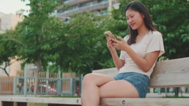 Şehir parkında bankta oturan mutlu genç kadın, cep telefonu kullanıyor.