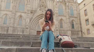 Genç bayan gezgin, eski kasaba meydanındaki merdivenlerde otururken cep telefonu kullanıyor.