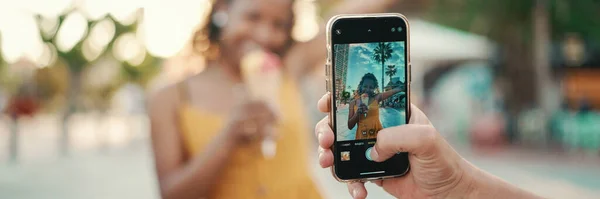 Close-up of a man taking a picture of a woman on a mobile phone. Closeup, smiling woman eating ice cream and filming it with smartphone on urban city background. Backlight