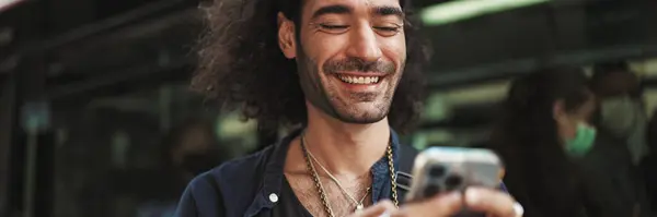 長い巻き毛と頑丈さを持つ若い魅力的なイタリア人男性は携帯電話を使用しています スマートフォンにメッセージを書くスタイリッシュな男 — ストック写真