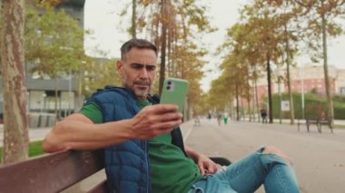 Orta yaşlı bir adam parkta bankta oturuyor ve cep telefonu kullanıyor.