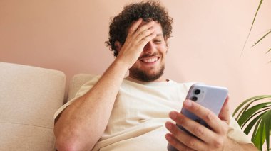 Yaklaş, kıvırcık saçlı ve sakallı, rahat oturma odasında koltukta oturan, cep telefonunu ekrana kaydıran genç adam.