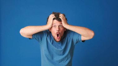 Mavi tişörtlü, kıvırcık saçlı bir adam kameraya büyük gözleriyle bakıyor ve ellerini kaldırıyor. Stüdyonun mavi arka planındaki iyi habere seviniyor.