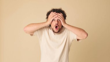 Bej tişörtlü heyecanlı adam kameraya bakıyor. Stüdyodaki bej arka planda şaşkınlıkla ellerini kaldırıyor.