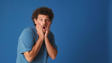 Mavi tişörtlü kıvırcık saçlı adam bir reklam, reklam panosu ya da stüdyonun mavi arka planında fotokopi alanı olan bir duyuru göstermek için başka bir yere bakıyor.