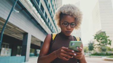 Gözlüklü, kahverengi elbiseli, sırtında sırt çantası olan genç bir kadın modern binalarda sokakta duruyor. Akıllı telefonunda harita uygulaması kullanıyor.