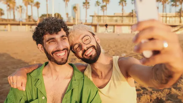 Genç eşcinsel çiftin sahilde otururken kucaklaşması, gülümsemesi ve cep telefonuyla selfie çekmesi.