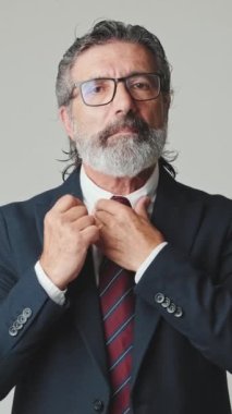 Orta yaşlı iş adamı gri arka planda izole edilmiş kameraya bakarken kravatını düzeltiyor.