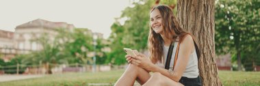 Beyaz bluzlu uzun dalgalı saçlı güzel kız parktaki ağacın altındaki yeşil çimlerin üzerinde otururken cep telefonu kullanıyor. Telefonundaki resimlere parmaklarınla bak.