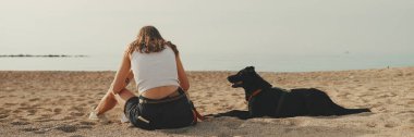 Beyaz bluzlu uzun dalgalı saçlı tatlı kız, çakıl taşı plajında siyah köpek deniz manzaralı oynuyor.