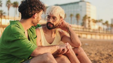 Birbirlerine aşık eşcinsel çift sahilde oturuyor, konuşuyor ve eğleniyorlar, kollarını kavuşturup sarılıyorlar.