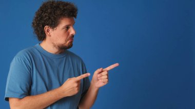 Kıvırcık saçlı, mavi tişörtlü, parmaklarıyla yer gösteren, reklam veren, reklam veren, reklam veren ya da stüdyodaki mavi arka planda duyuru yapan adam.