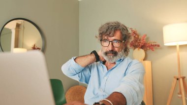 Gri saçlı, gözlüklü, yorgun yaşlı bir adam modern dairede oturma odasında oturmuş dizüstü bilgisayarla çalışıyor.