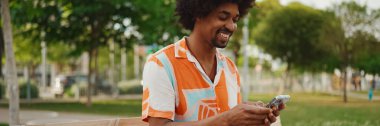 Parktaki bankta oturan Afro-Amerikan gömlekli genç adam akıllı telefonunu kullanıyor. Gülümseyen Afro-Amerikan bir adamın cep telefonundaki fotoğraf videosuna yakından bakması. Yaşam tarzı kavramı