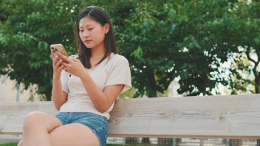 Şehir parkında bankta oturan güzel esmer kız, cep telefonu uygulaması kullanıyor, sosyal medyada sörf yapıyor, haberleri kontrol ediyor.