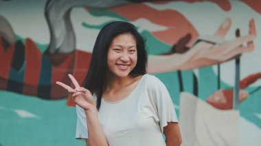 Güzel, mutlu esmer kız, günlük giysiler içinde, kameraya bakıyor, poz veriyor, zafer işaretini parmaklarıyla gösteriyor.