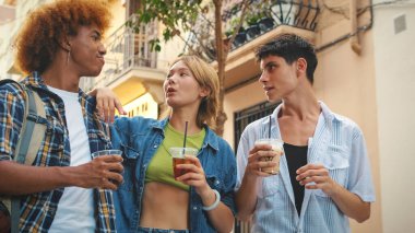 Bir grup mutlu arkadaş, iki erkek ve bir kız, ellerinde içkilerle şehir caddesinde yürüyor, sohbet ediyor ve gülüyorlar.