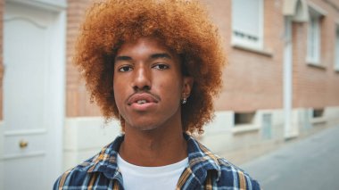 Afrikalı saçlı, mavi gömlekli, gülümseyen adam sokakta dikilirken kameraya bakıyor.