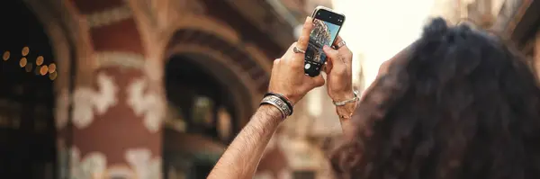 Young attractive italian guy with long curly hair and stubble takes photo of an old building. Stylish man with an earring in his ear and lot of chains photographing sights.