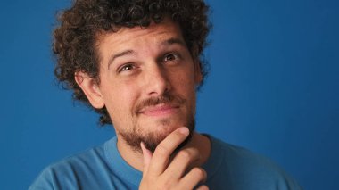 Yakından bakınca, mavi tişörtlü kıvırcık saçlı adam zor bir seçim düşünüyor, aklında bir plan kuruyor, stüdyoda mavi arka planda izole edilmiş bir plan.