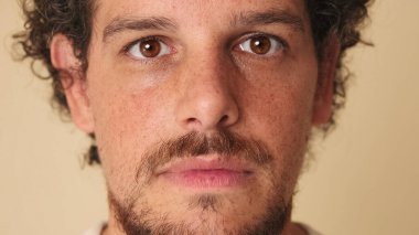 Kıvırcık saçlı ve çilli bir adamın yakın plan portresi. Stüdyoda bej arka planda kameraya bakıyor.